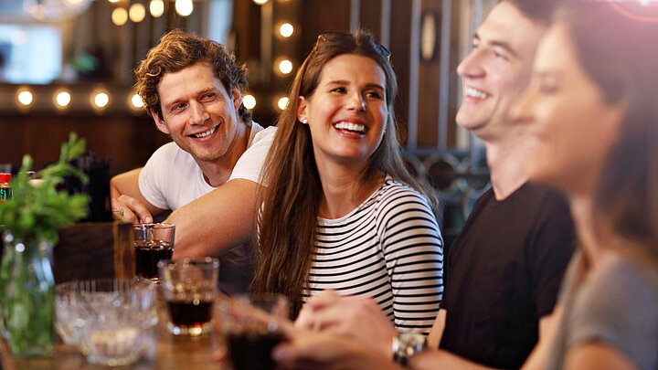 Grafika dekoracyjna. Cztery uśmiechnięte osoby siedzą obok siebie przy długim stole w restauracji. Przed sobą mają szklanki z napojami, śmieją się i rozmawiają. 